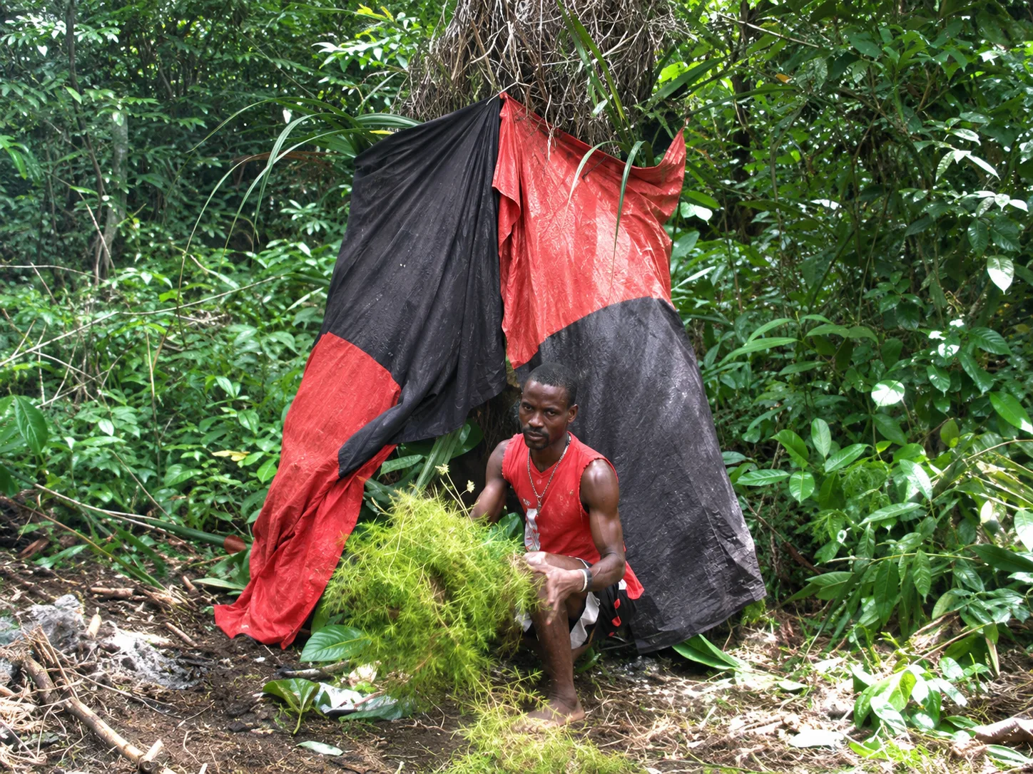 Un officiant assis devant un abri rituel rouge et noir, en pleine forêt — la tradition transmise au quotidien